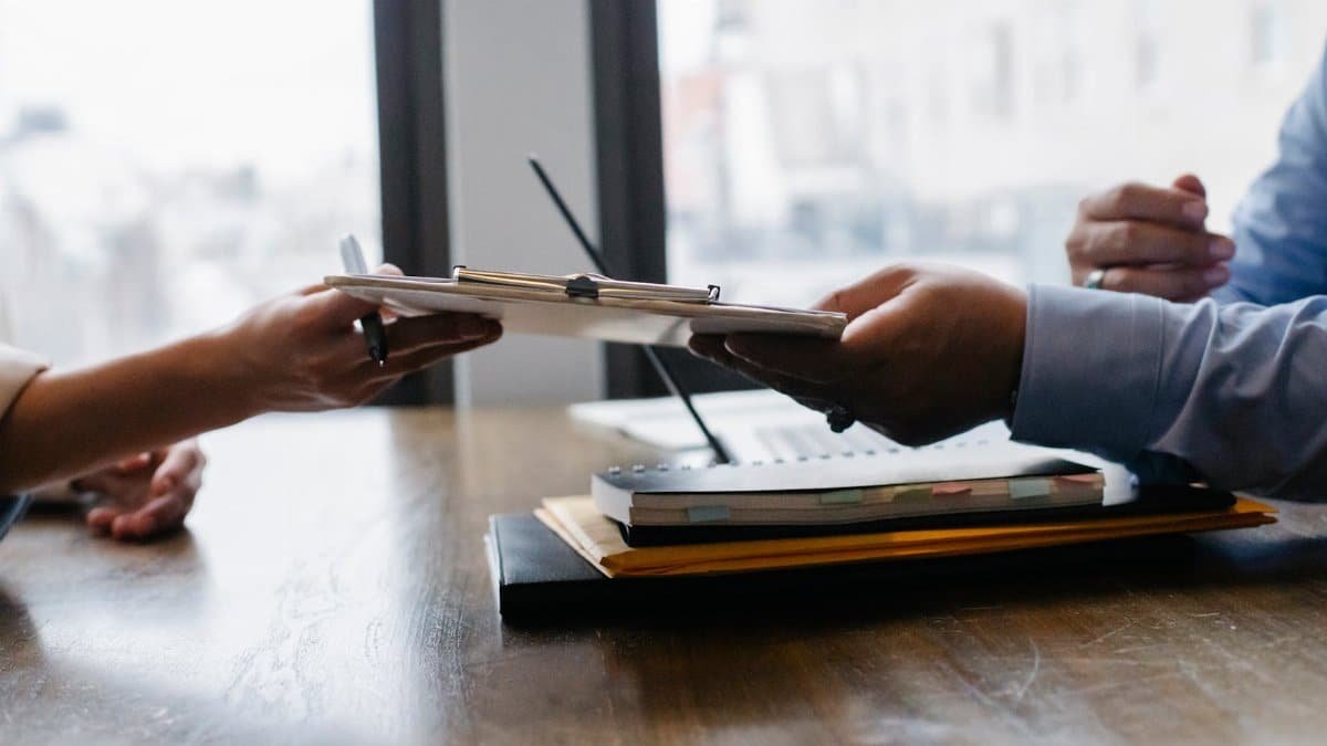 Crop anonymous ethnic woman passing clipboard to office worker with laptop during job interview