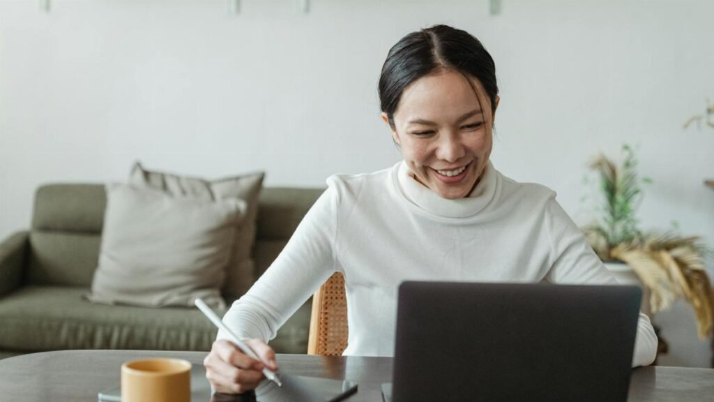 A cheerful woman uses a laptop and tablet for a video call, working remotely in a cozy living room.