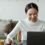 A cheerful woman uses a laptop and tablet for a video call, working remotely in a cozy living room.