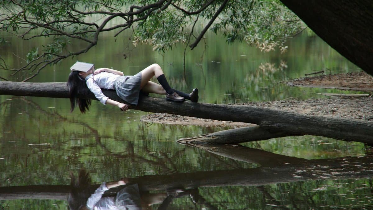 A young woman with a book on her face lies relaxing on a tree trunk by a tranquil lake.