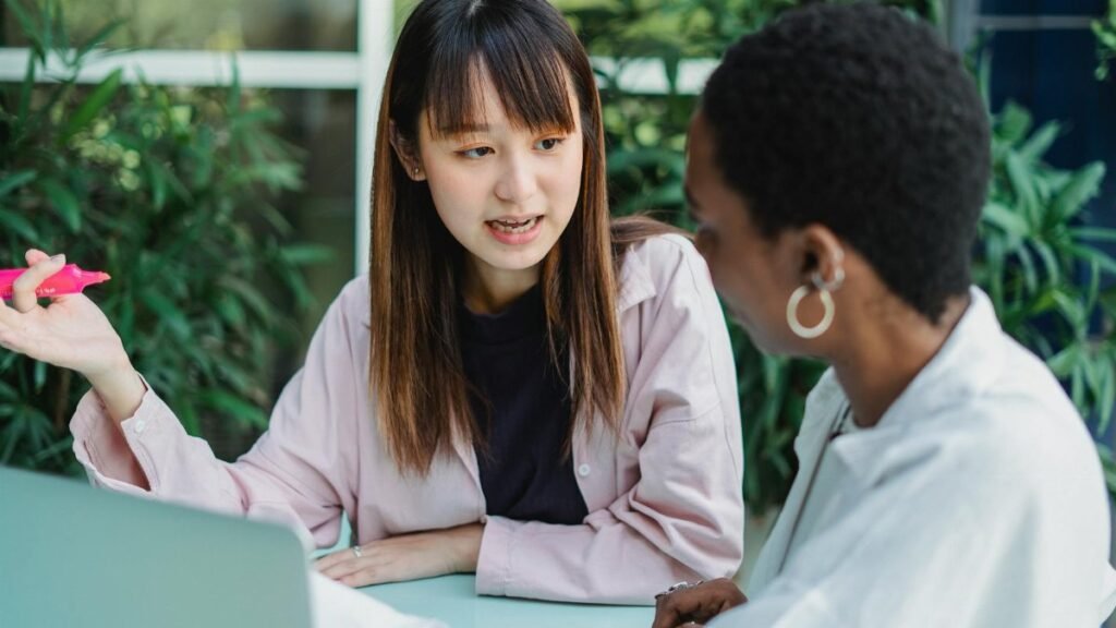 Young attentive ethnic female with marker conversing with African American partner at table with laptop while looking at each other