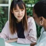 Young attentive ethnic female with marker conversing with African American partner at table with laptop while looking at each other