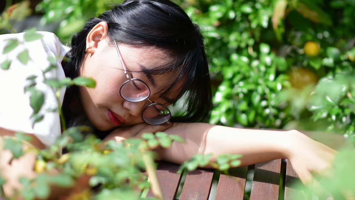 A young woman with glasses relaxes on a wooden table in a lush garden setting, surrounded by greenery.