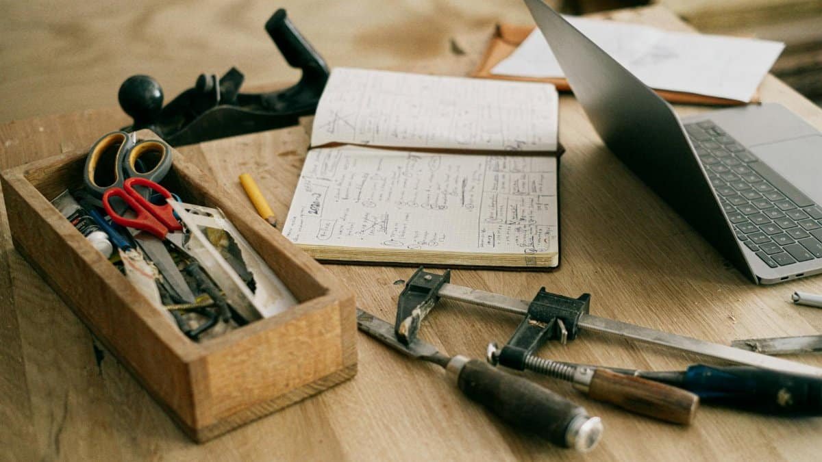 A creative woodworking workspace with tools, notebook, and laptop on a wooden table.