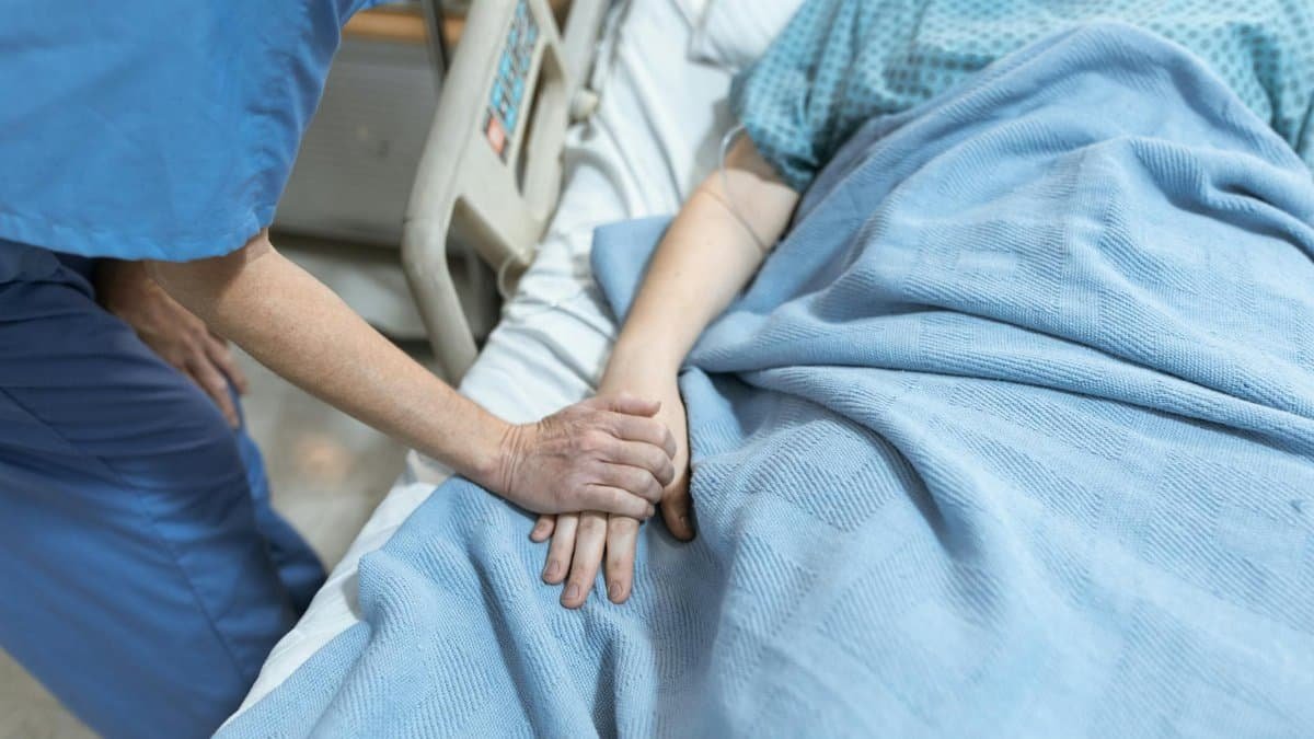 A nurse in scrubs offers comfort by holding a patient's hand in a hospital bed.