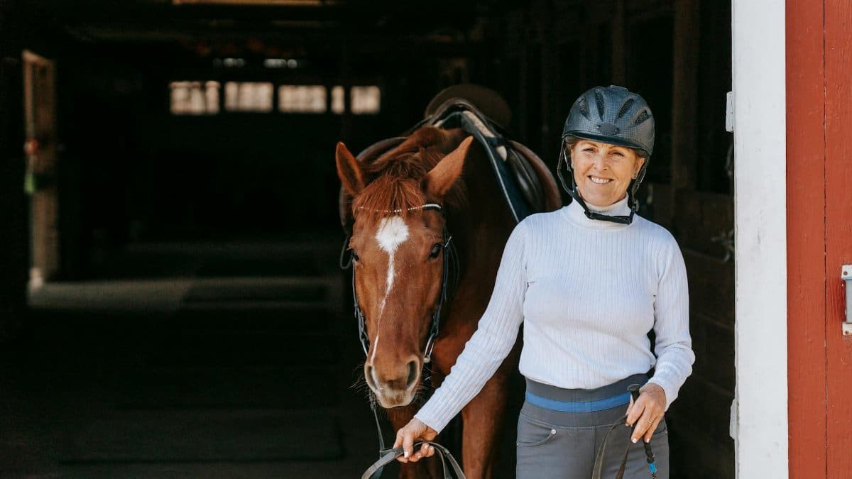 A smiling woman in riding gear leading a saddled horse outdoors.