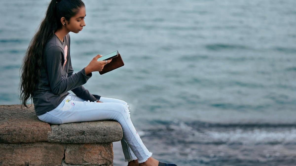 A young woman enjoys a peaceful moment by the sea, engrossed in her smartphone at Diu, India.