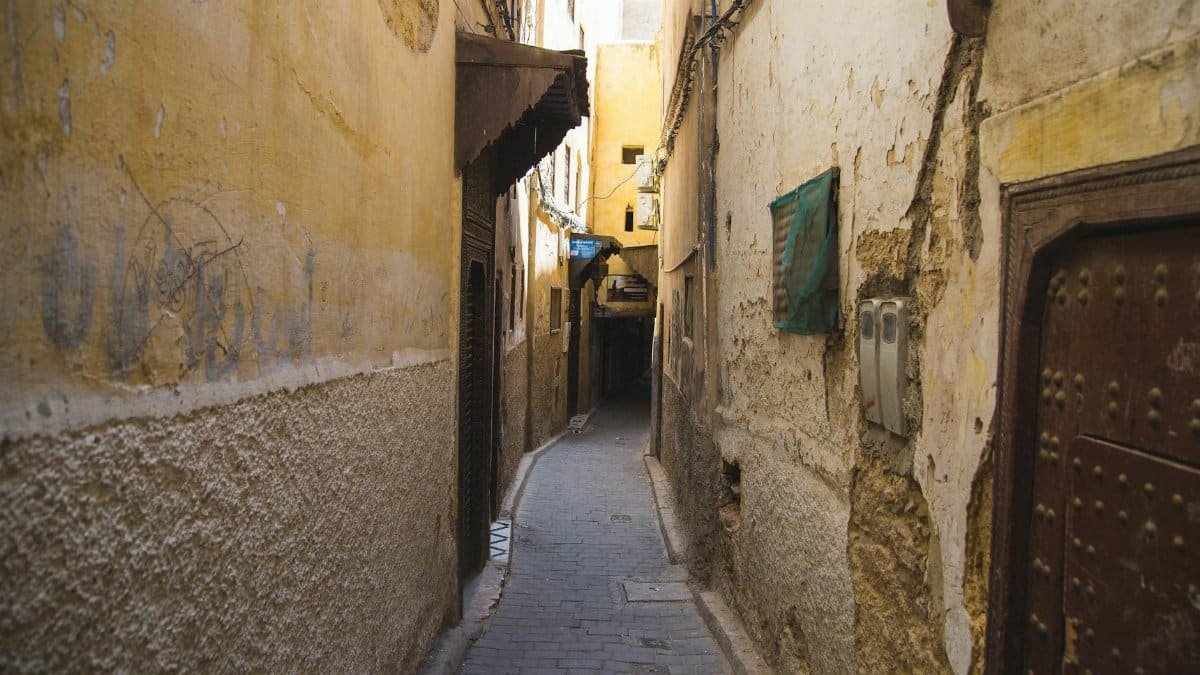 Paved narrow pedestrian walkway between weathered stone residential buildings with metal doors in old Eastern town on sunny weather