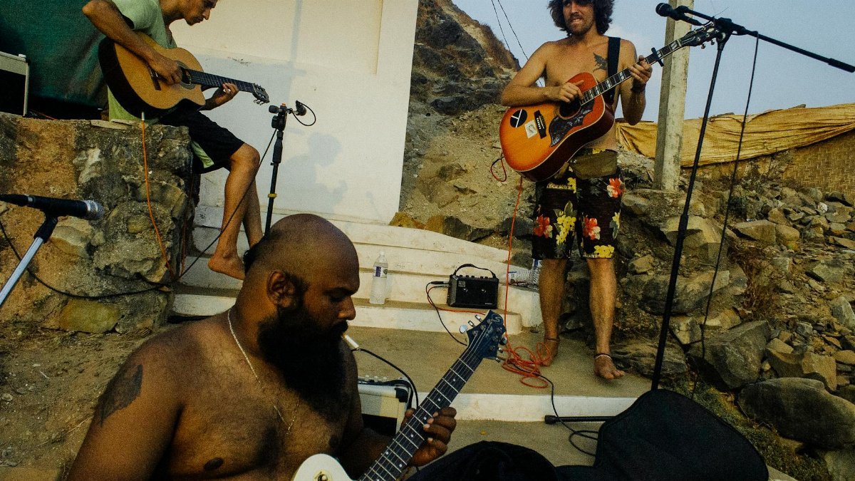 Local musical band of ethnic males in casual wear playing guitars outside shabby house in rural location