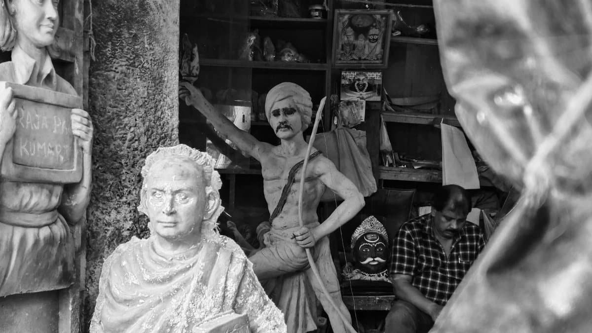 Black and white photo of sculptures and artisan in Kumartuli, Kolkata showcasing cultural craftsmanship.