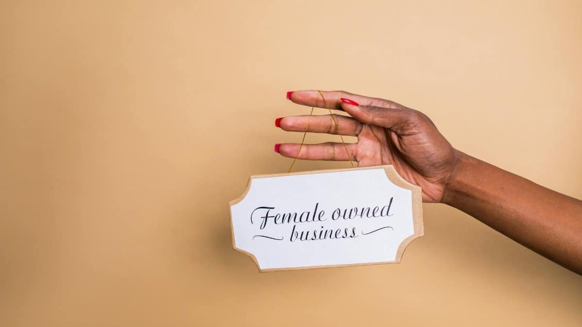 Close-up of a woman's hand holding a 'Female Owned Business' sign on a beige background.