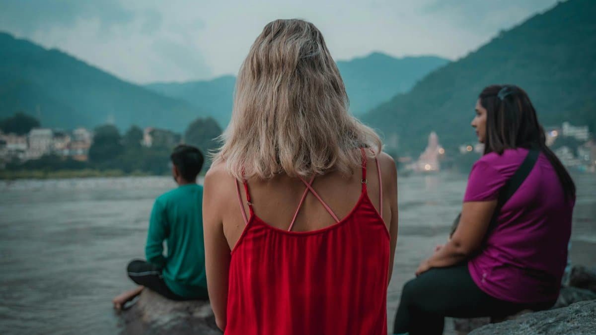 Three people meditate by the Ganges River in Rishikesh, India, with a serene mountain backdrop.