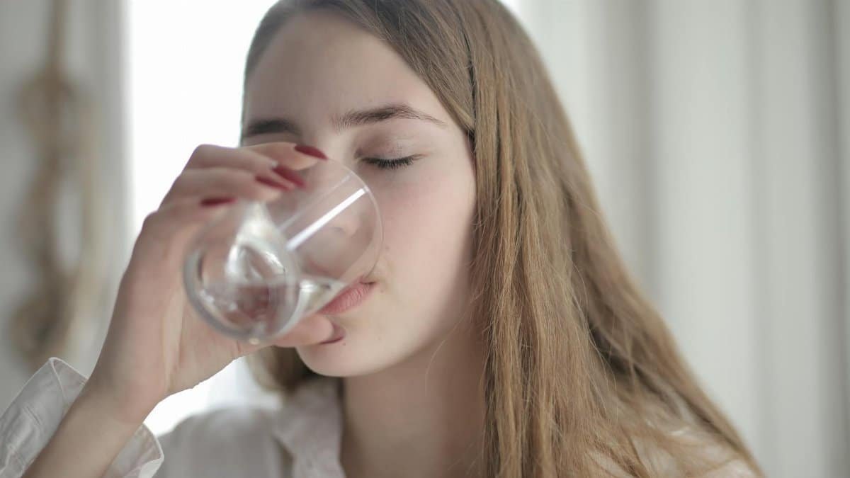 A young woman with brown hair drinks water from a clear glass indoors, eyes closed.