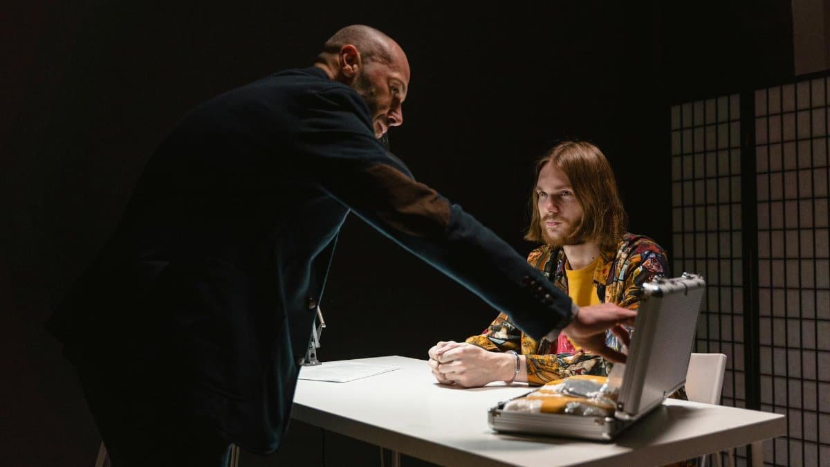 Dramatic interrogation scene with two men discussing a briefcase inside a dark room.