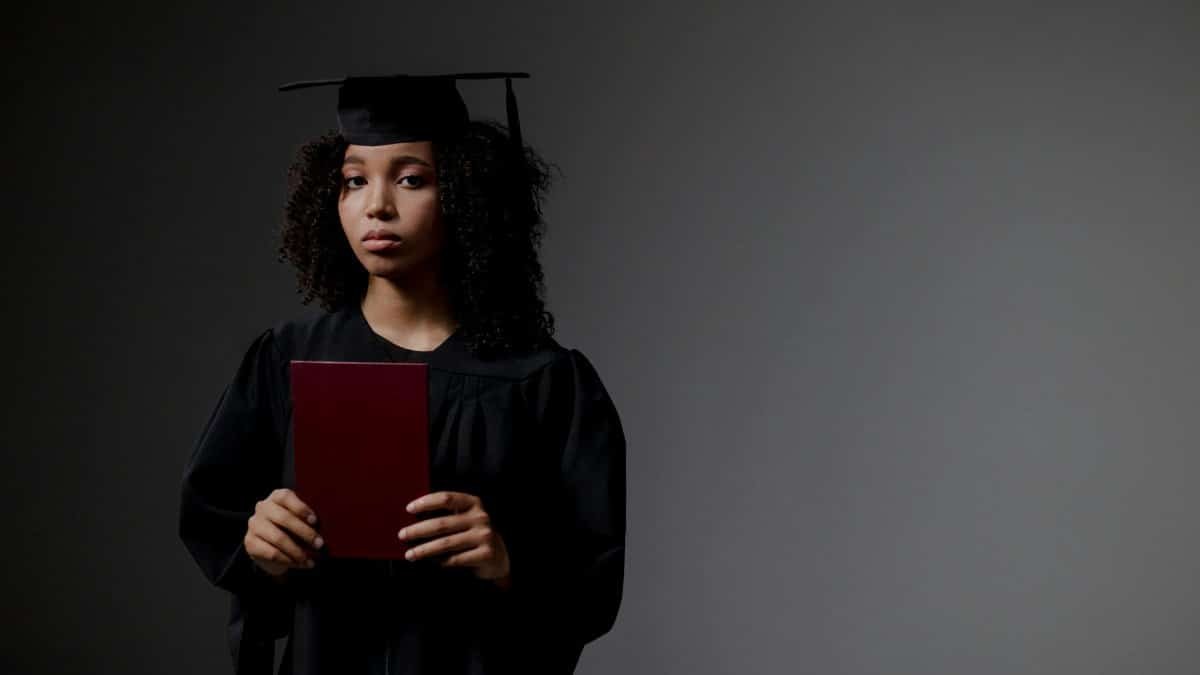 Young graduate in black academic regalia holds diploma, looking serious.