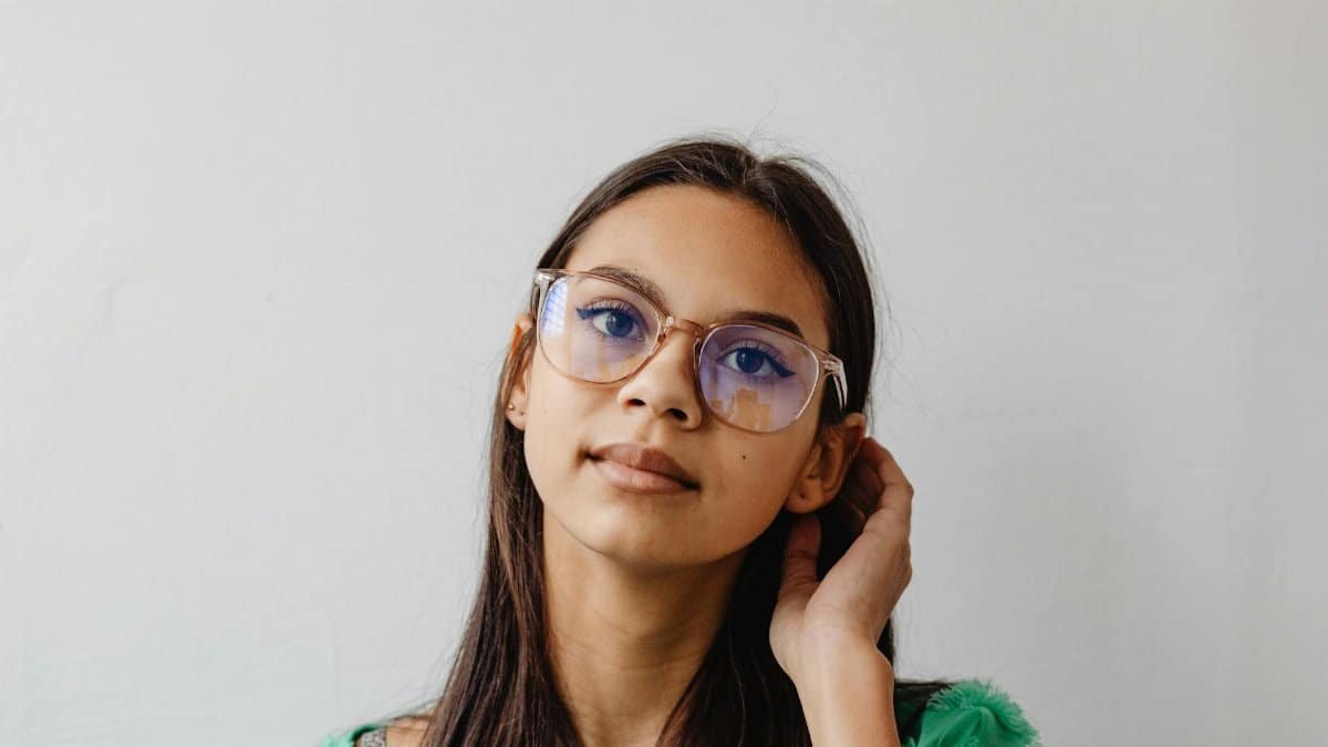 A thoughtful young woman wearing eyeglasses, posing indoors against a light background.