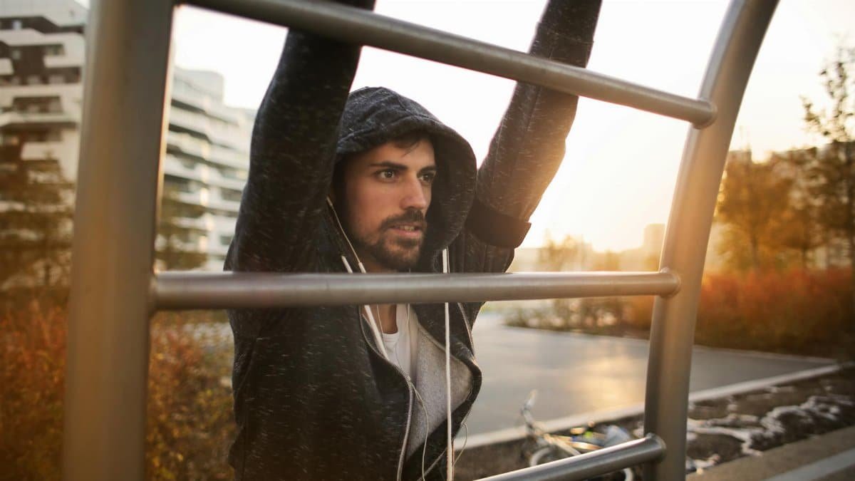 Young athletic male in sportswear with earphones doing exercises on ladder while training on sport ground near river in autumn morning in city