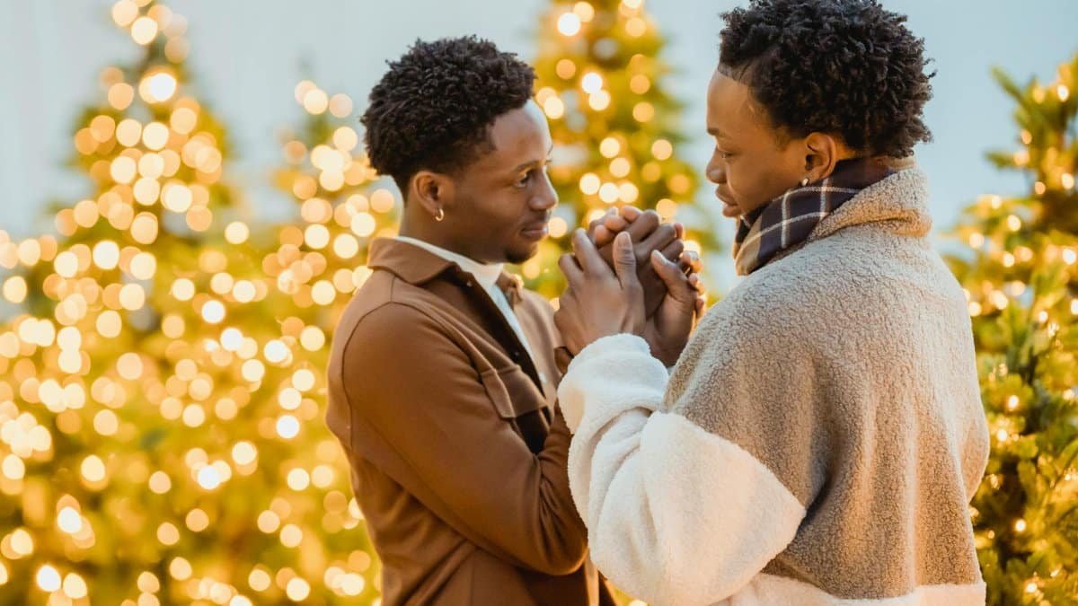 Side view of romantic African American homosexual couple holding hands and looking at each other while standing near glowing garlands on blurred background