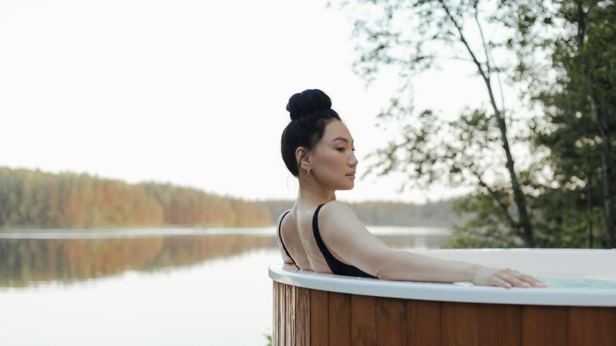 Asian woman enjoying a serene moment in a hot tub overlooking a tranquil lake.