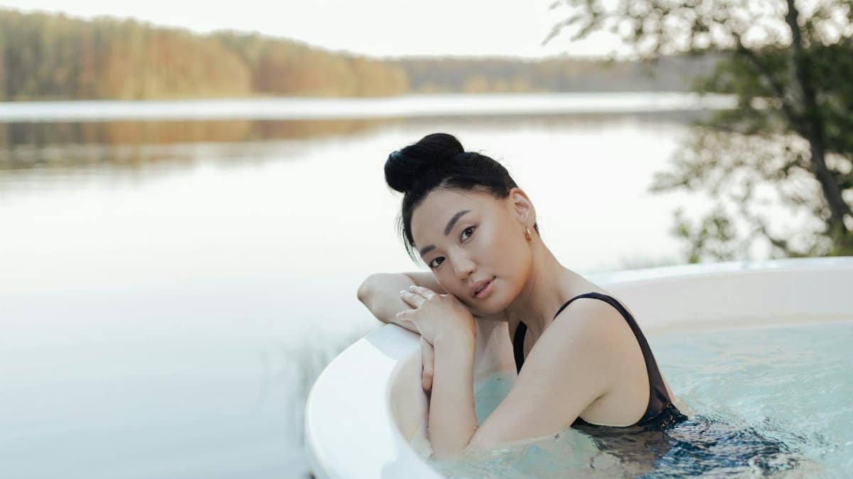 A woman enjoys a relaxing time in an outdoor hot tub by a serene lakeside setting.