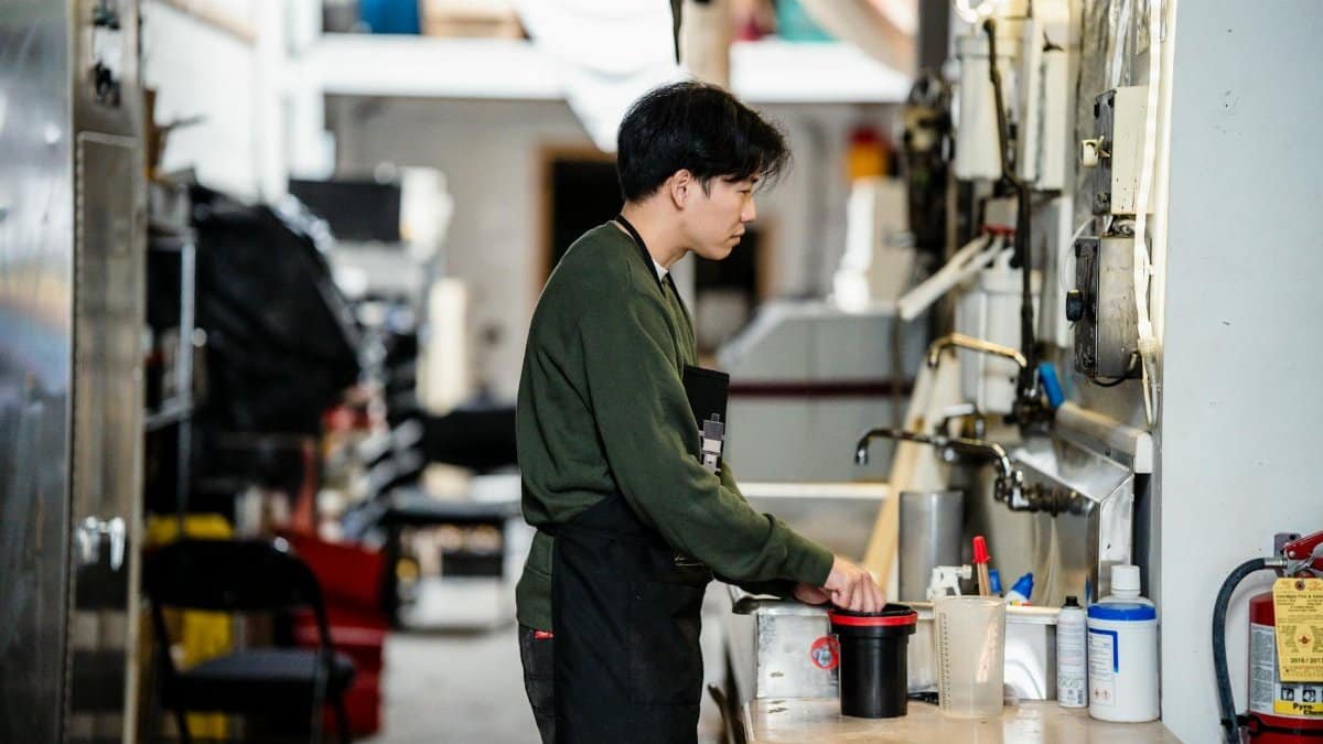 Asian man in green sweater mixing chemicals in an industrial workspace indoors, focused on work.