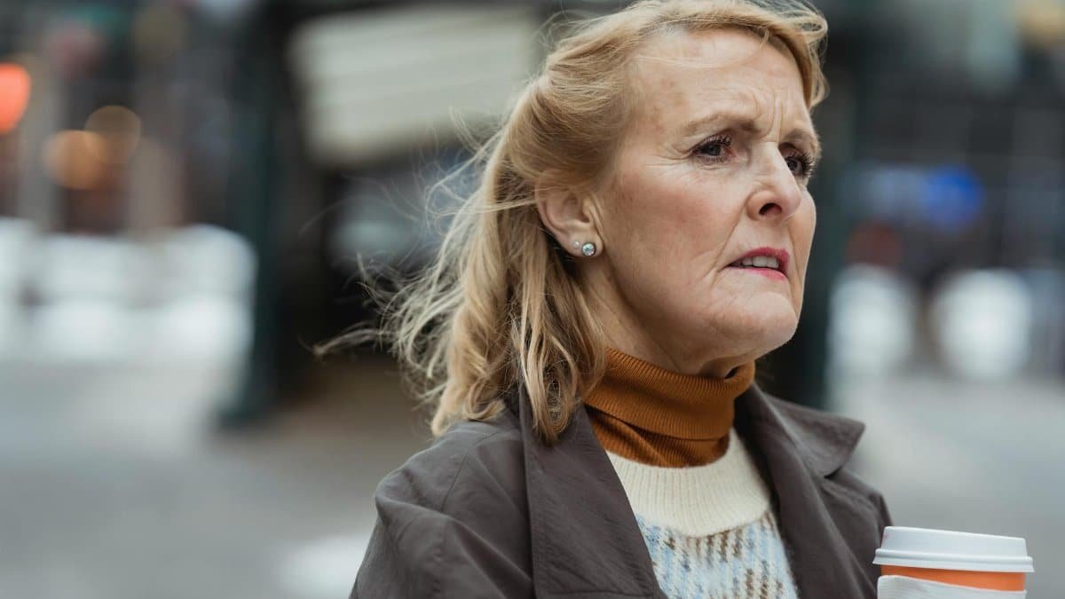 Pensive elderly female with takeaway hot drink looking away in town on windy day on blurred background