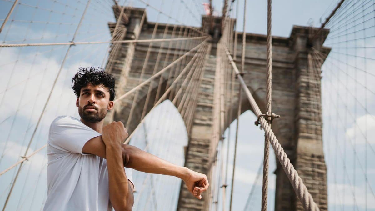 Man in white shirt stretching on Brooklyn Bridge, showcasing fitness and wellness in a city setting.