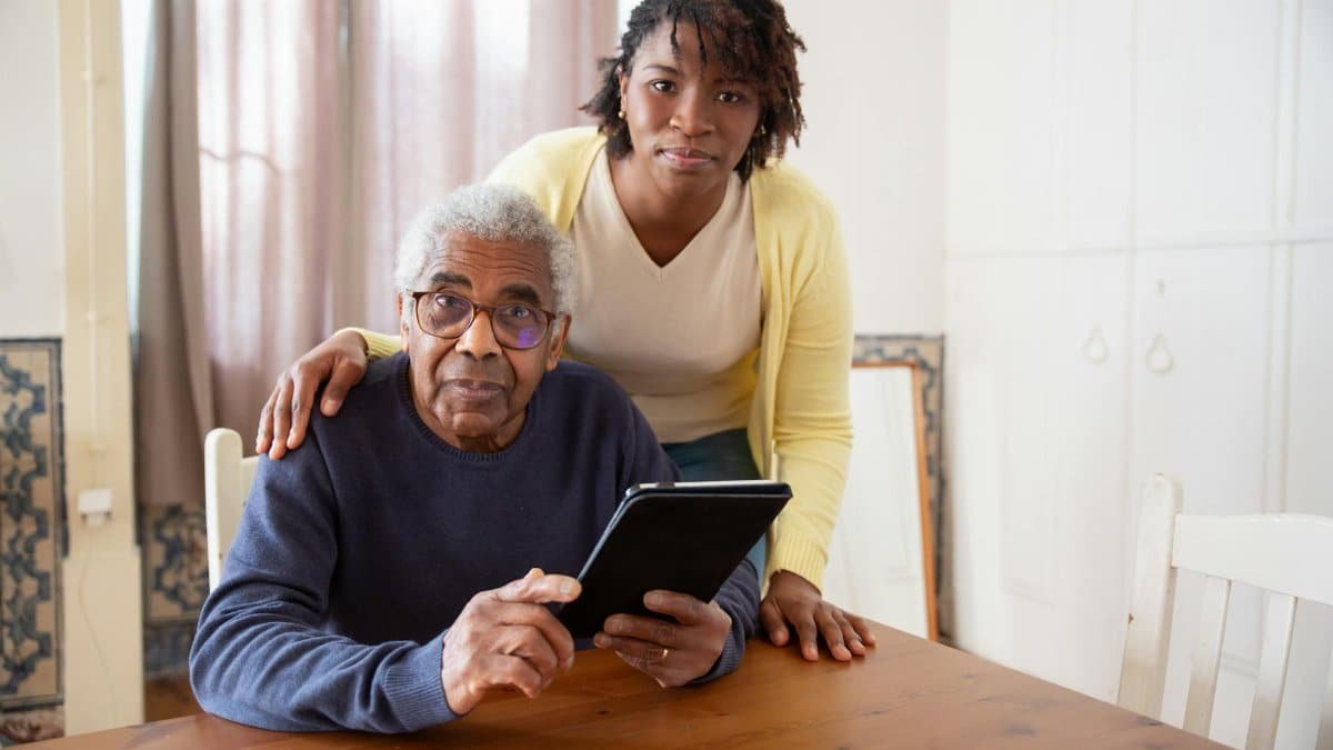 A senior man with eyeglasses and caregiver using a tablet at home, focusing on care and companionship.