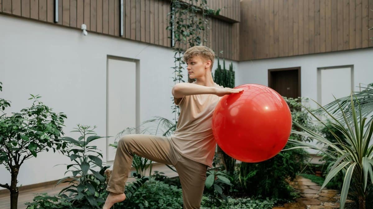 Adult male practicing balance exercise with a red balloon in a greenhouse setting.