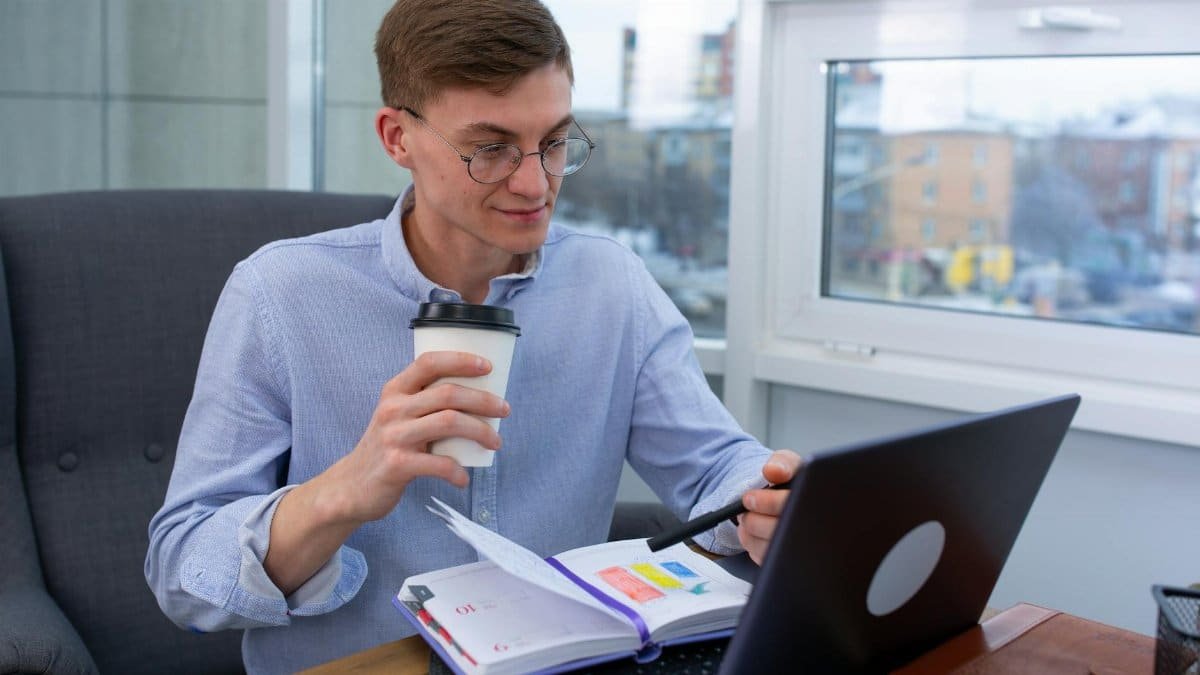 Business professional sipping coffee while working on a laptop with a planner at an office.