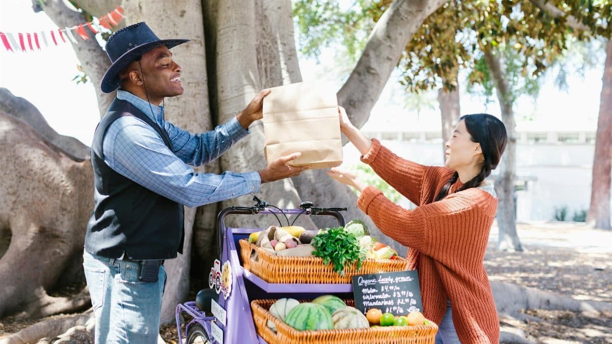 A vendor hands a paper bag to a customer at an outdoor farmer's market filled with fresh produce.