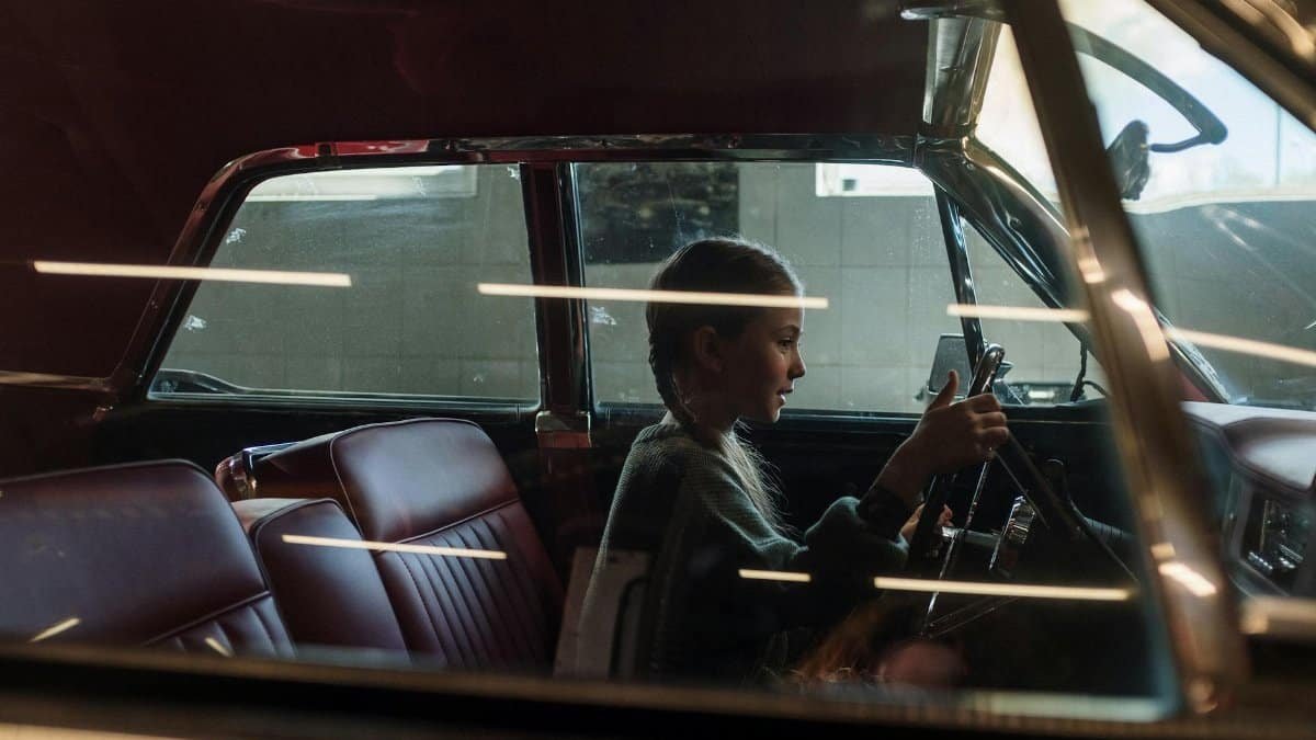 A young girl with pigtails sits in a vintage car, holding the steering wheel with focus.