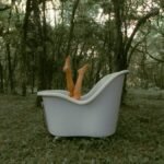 Unrecognizable female lying in bathtub with legs raised on grassy terrain near tall lush trees in woodland during daily routine