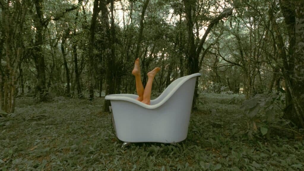 Unrecognizable female lying in bathtub with legs raised on grassy terrain near tall lush trees in woodland during daily routine
