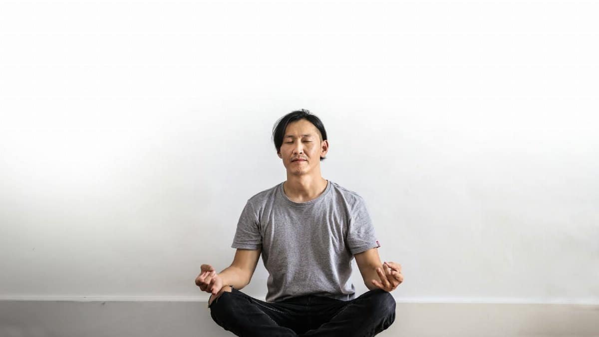 Asian man practicing meditation indoors on a wooden floor, promoting relaxation and mindfulness.