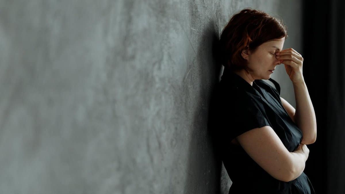 A woman in stress with eyes closed, leaning against a concrete wall indoors.