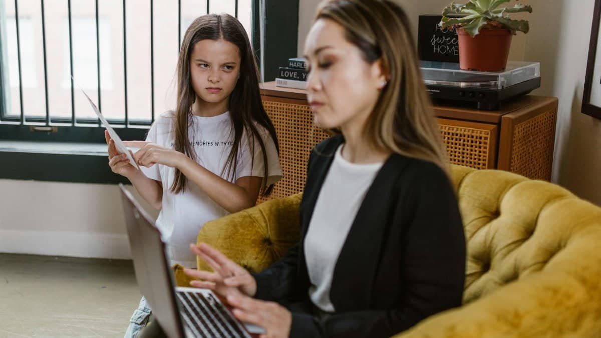A woman in a black blazer uses a laptop on a couch while a young girl stands near holding paper.