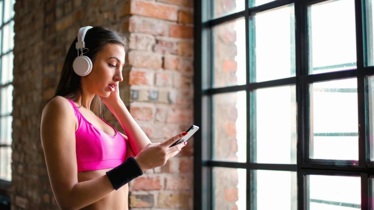 Woman in sports wear listening to music on headphones while holding a smartphone indoors.