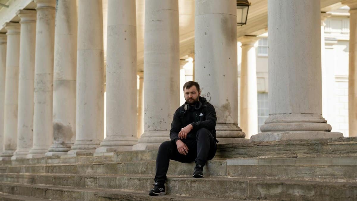 A man in black clothing sits on stone steps among large columns in daylight.