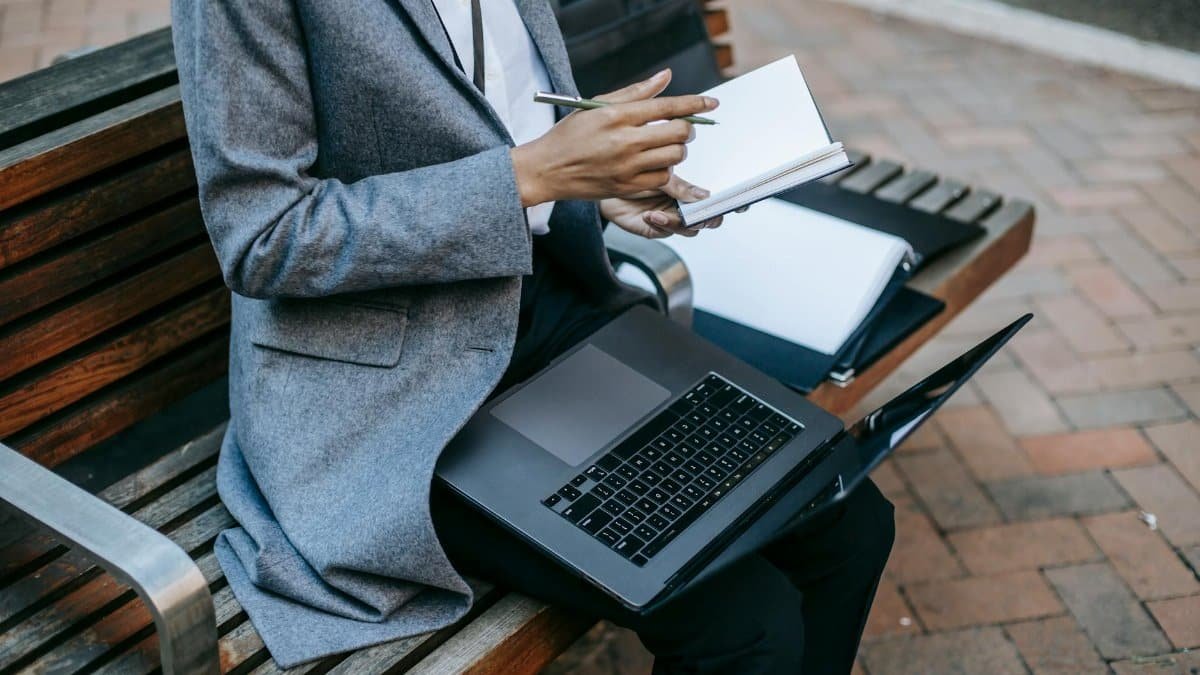Businesswoman taking notes and using a laptop on a bench outdoors.