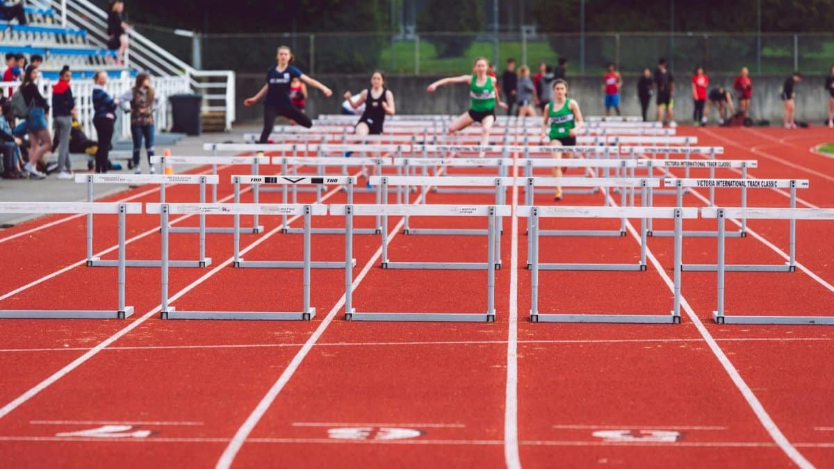 Women athletes compete in a hurdles race at a Victoria track event on a sunny day.