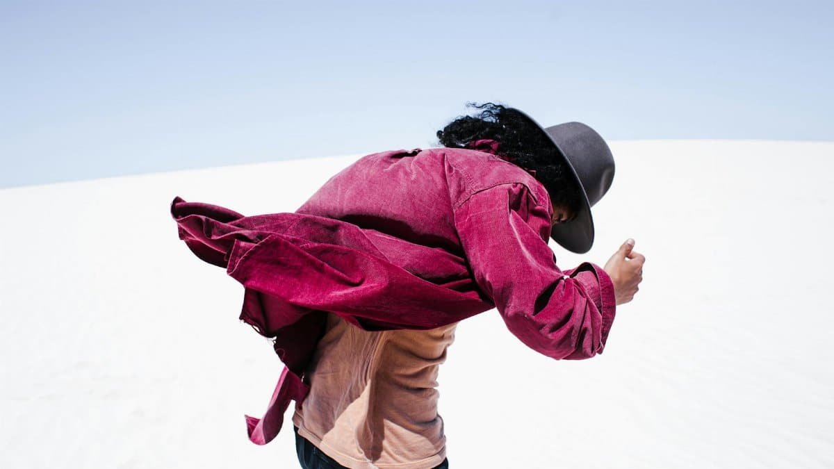 A person with pink sleeves and a fedora bravely faces strong wind in a vast desert.