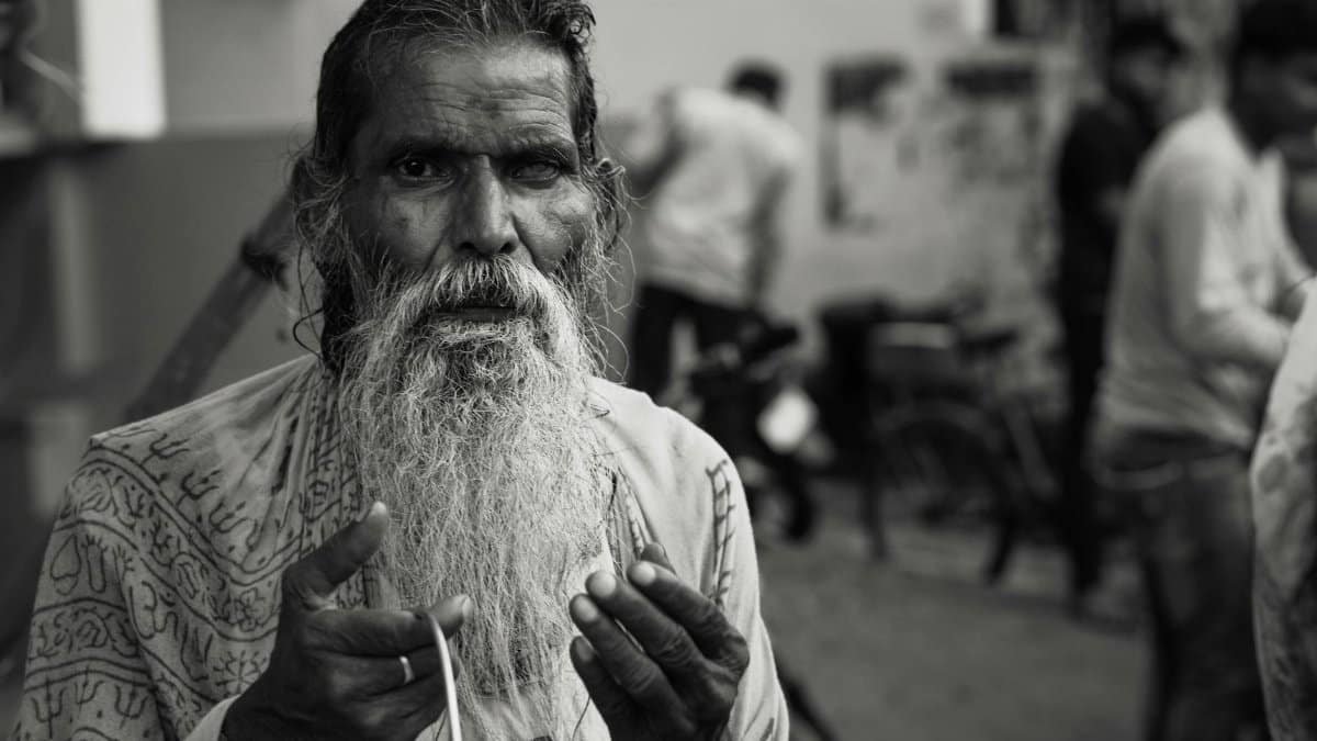 A senior man with a long beard sits on a city street in Varanasi, India, seeking help.