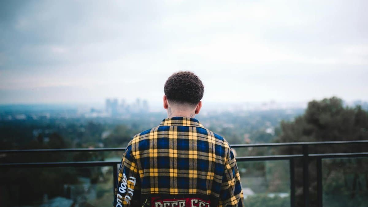 Man in plaid shirt stands on balcony with blurred city view in background.