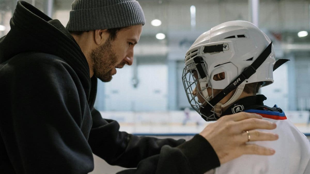 A father guiding and encouraging his son in full ice hockey gear at an indoor rink.