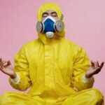 Man in protective suit and respirator practicing meditation against pink backdrop.