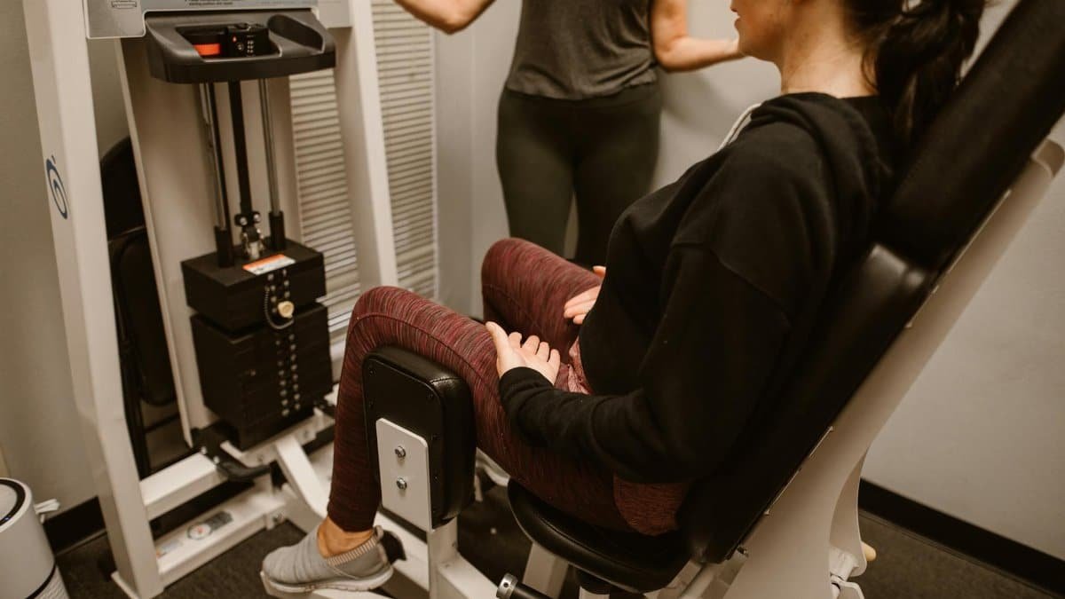 A woman performs exercises on a hip abductor machine with guidance from a trainer.