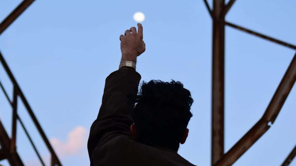 A man silhouetted against the morning sky reaches towards the moon with an outstretched hand.