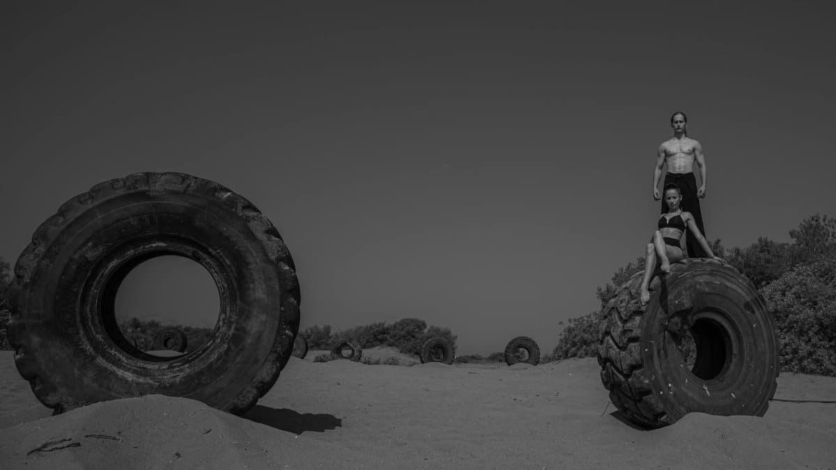 Strong athletic man and woman exercising on large tires in a sandy outdoor setting.