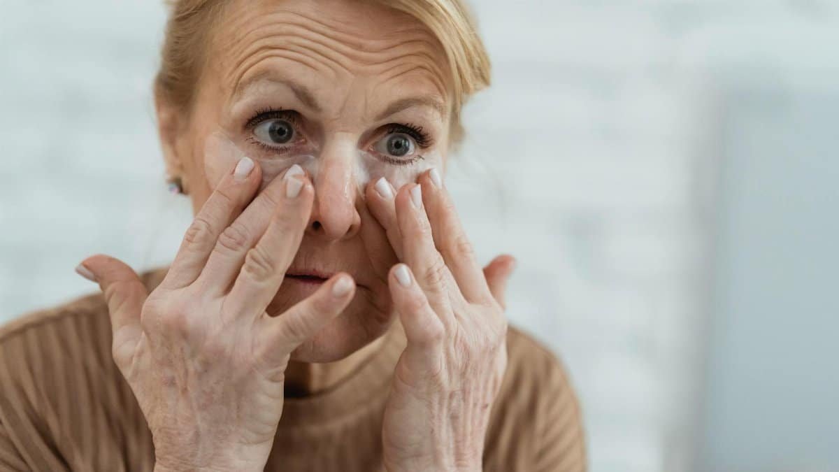 Crop focused mature female applying moisturizing nourishing eye patches and looking away in light bathroom